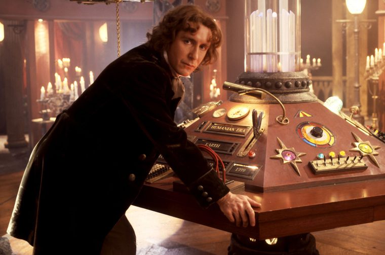 The image shows a man in his forties in a dark Victorian-style coat leaning over an elaborate, hexagonal console covered in various dials, switches, levers, and decorative elements. The console has labels visible including "HUMANIAN ERA," "EARTH," and "1999." Rising from the center is a tall cylindrical glass column. The atmospheric set is lit by candlelight in the background, giving it a warm, gothic feel — very much the interior of a TARDIS console room!