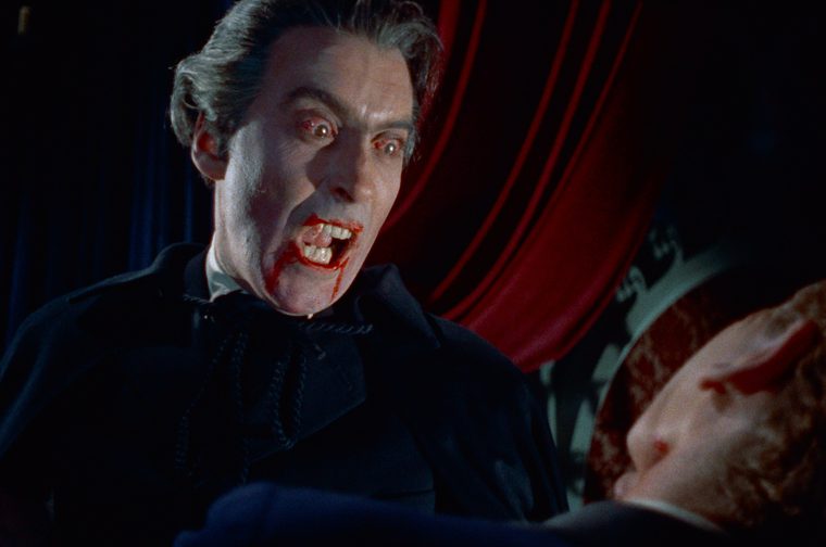 A pale, dark-haired vampire actor is mid-snarl, fanged mouth smeared with blood, eyes bloodshot and wide with fury, wearing a black high-collared cape, with a red-curtained set behind him and another actor's blood-streaked face in the foreground.