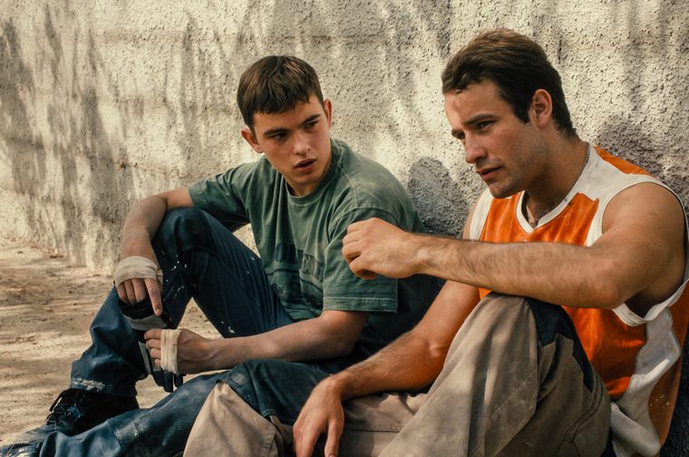 Two young men are sitting against a concrete wall outdoors. The one on the left wears a faded green t-shirt and jeans, with bandaged hands, and appears to be speaking. The one on the right wears an orange and white sleeveless top and light-coloured trousers, his fist raised, looking pensive. The scene has a gritty, naturalistic feel.