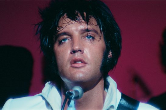 A close-up image of Elvis Presley during his Las Vegas residency, with long sideburns and a sweaty head of hair. A microphone is in front of his mouth and we can see shadows of band members behind him in front of a dark red backdrop.
