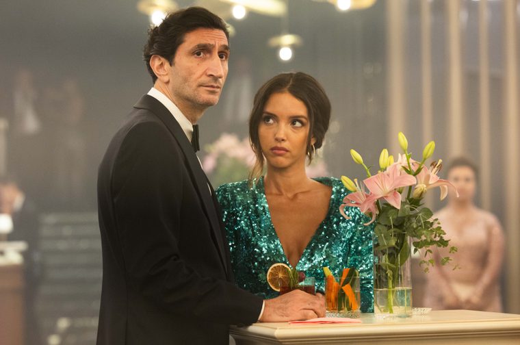 Two formally dressed people stand at a bar or high table in an elegant, dimly lit venue. The man on the left wears a black tuxedo and looks upward with a serious expression. The woman beside him wears a sparkling teal/emerald sequined dress and glances sideways with a wary look. On the table in front of them are cocktails and a glass vase with pink lilies. Other guests are visible in the blurred background.