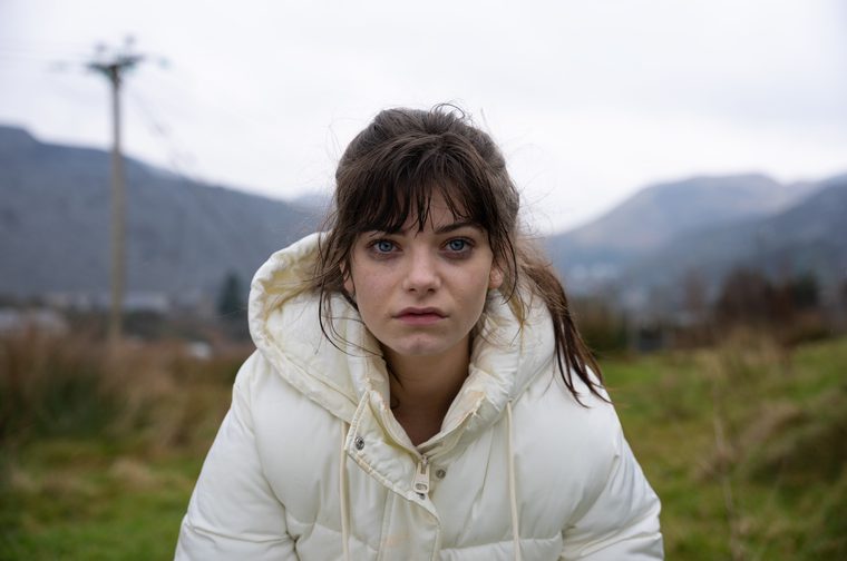 A young woman with dark brown hair and striking blue eyes leans forward, gazing intensely at the camera. She wears a cream-coloured puffer jacket with the hood up around her shoulders. Her hair is slightly dishevelled, with a fringe across her forehead. The background shows an overcast rural landscape with rolling hills, dry grass, and a telegraph pole, suggesting a cold, grey day.
