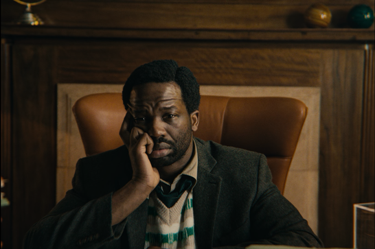 A man with short dark hair and a beard sits in a tan leather wingback chair in a wood-paneled office, resting his cheek on his hand with a weary, thoughtful expression. He's dressed in a green-and-cream striped sweater vest, tie, and dark tweed blazer. The room has a warm, vintage feel, with decorative objects lining a shelf in the background.