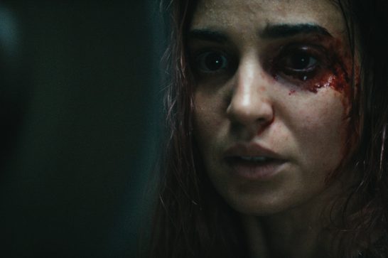 A close-up still of a young woman with long dark hair, her face partially illuminated against a dark, shadowy background. She has a bloody wound near her right eye, with streaks of blood running down her cheek. Her expression is wide-eyed and fearful, mouth slightly open. The overall tone is dark and atmospheric, consistent with horror film cinematography.