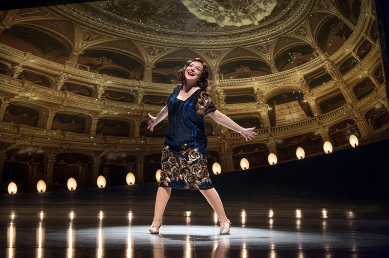 A female performer stands centre stage on a brightly lit theatre stage, arms outstretched and smiling, wearing a blue and gold sequined flapper-style dress with gold floral embroidery and silver T-strap heels. The stage is lined with warm amber footlights. Behind her, a grand ornate opera house interior is projected as a backdrop, featuring gilded balconies, arched ceilings with painted murals, and warm glowing wall sconces.