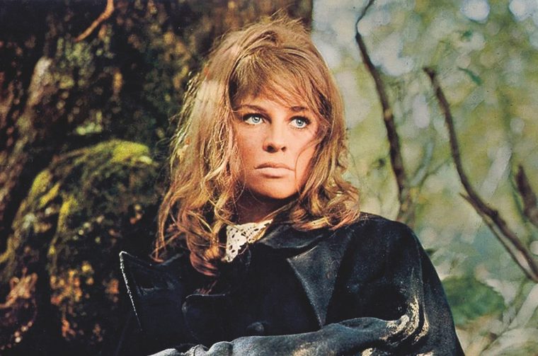 A younger woman (Julie Christie) with tousled blonde hair and blue eyes, dressed in a dark Victorian-era cloak with a lace collar, stands against a moss-covered tree in a woodland setting. The image has a warm, vintage film quality.