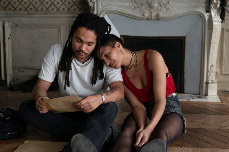 Two young adults sit together on a wooden parquet floor in front of an ornate white fireplace, reading a letter or document. The male on the left wears a white t-shirt and has long dreadlocks; the female on the right leans on the man's shoulder, wearing a red tank top and denim skirt. The setting appears to be an elegant but bare room with decorative wallpaper.