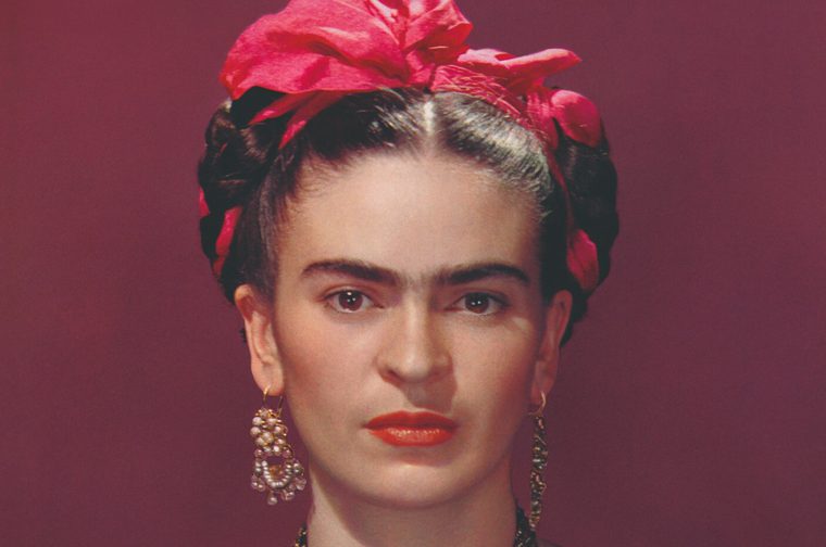 A portrait photograph of artist Frida Kahlo - dark braided hair adorned with a large red flower, elaborate pearl and gold drop earrings, bold red lipstick, and prominent natural eyebrows. The photo has a rich crimson/burgundy background.