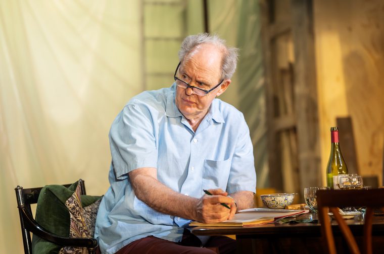 A grey-haired man in glasses and a light blue short-sleeved shirt sits at a table, pen in hand over an open notebook, looking off to the side with a thoughtful, slightly wary expression. The table holds a wine bottle, glasses, and a patterned bowl, set against a softly lit backdrop of draped fabric and wooden beams.
