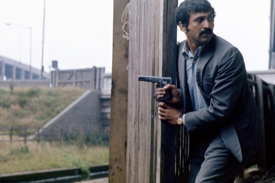 A middle aged South Asian man with a mustache, wearing a grey suit and gold watch, crouches behind a wooden post while holding a handgun raised near his shoulder. He peers cautiously around the post toward something off-frame. The setting appears to be an industrial waterside area with a canal lock, motorway overpass, and metal railings in the background.