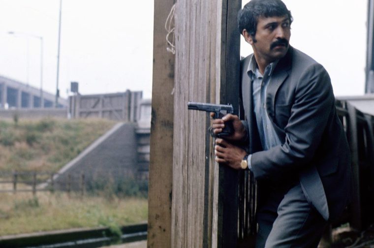 A middle aged South Asian man with a mustache, wearing a grey suit and gold watch, crouches behind a wooden post while holding a handgun raised near his shoulder. He peers cautiously around the post toward something off-frame. The setting appears to be an industrial waterside area with a canal lock, motorway overpass, and metal railings in the background.