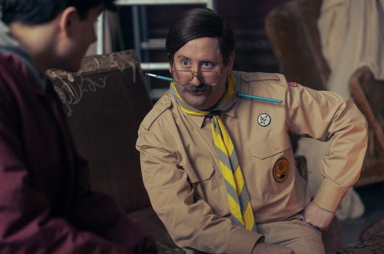 Image of a moustached man with slicked-down dark hair, wearing gold-rimmed aviator glasses perched low on his nose, a tan scout-style shirt with badges, and a yellow and grey striped neckerchief. A blue arrow-like pencil appears to pass through his neck. He crouches with one hand on his hip, staring with a raised eyebrow at a blurred, dark-haired figure in a maroon jacket in the foreground. They are in a cluttered room with worn armchairs and sheet-draped furniture.