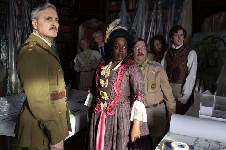 Image of a group in assorted historical costumes gathered in a dark library with bookshelves and hanging plastic sheeting. In the foreground, a grey-haired man with a mustache wears a olive-green military officer's uniform with a leather belt. Beside him, a woman in a mauve brocade gown with ruffled pink and red trim, yellow bows and a tall white feather in her hair looks sideways. Behind her, a mustached man in a tan scout uniform and aviator glasses, a shaggy-haired figure with a beard, and a man in a patterned waistcoat and teal cravat all stare in the same direction. Two more figures, a woman in an Edwardian-style blouse and a man in a suit, look upward in the background. Architectural plans, a white scale model of a building and a roll of masking tape sit on tables around them.