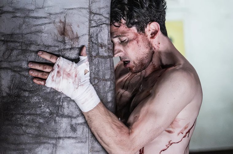 A shirtless, bloodied young man leans exhausted against a large leather punching bag, resting his forehead against it with eyes closed and mouth open, breathing heavily. His hand is wrapped in a white boxing hand wrap stained with blood, gripping the edge of the bag. Streaks of blood run down his cheek, neck, chest, and torso. The background is softly blurred, suggesting a dim gym or boxing training space.
