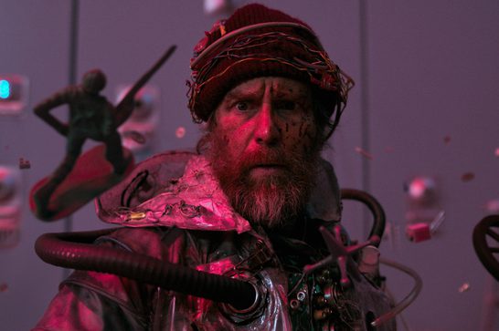 A bearded, scruffy man in his fifties wearing a ragged beanie hat stares with an incredulous expression at the viewer. He is wearing a shiny silver suit with black pipes sticking out of it and various random objects are floating around him, including a small wheel and a toy soldier.
