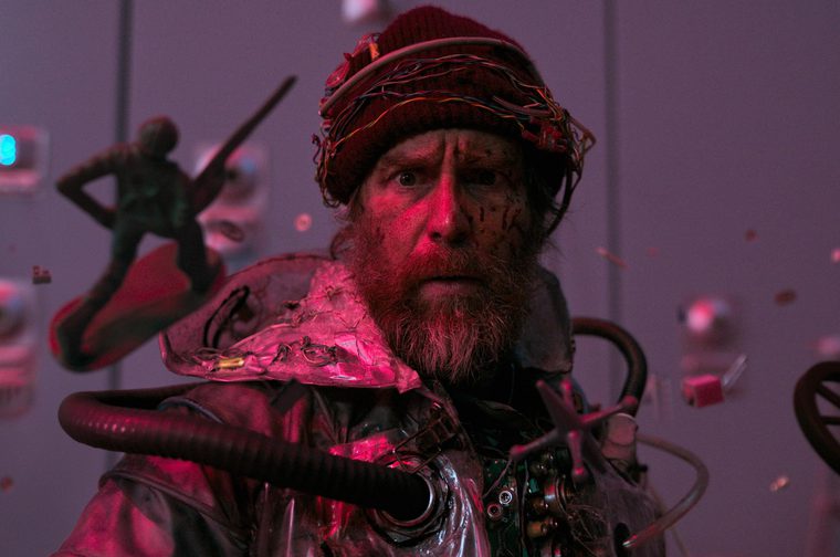 A bearded, scruffy man in his fifties wearing a ragged beanie hat stares with an incredulous expression at the viewer. He is wearing a shiny silver suit with black pipes sticking out of it and various random objects are floating around him, including a small wheel and a toy soldier.