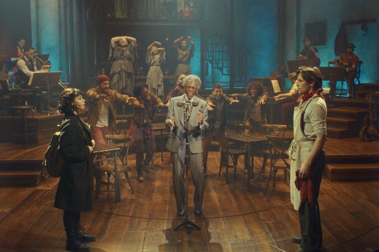Production photo from the musical Hadestown, showing the full ensemble on a wooden stage set designed like a tavern, with a live band positioned on platforms to either side. In the center, Hermes sings into a vintage microphone wearing a light gray suit, gesturing toward the audience. To his left, Eurydice stands alone in a black coat with a backpack, looking toward him. To his right, Orpheus stands in a cream-colored work shirt and apron with a red cloth in his hand, gazing back at Eurydice across the circle. Behind Hermes, the ensemble of workers reaches out with extended arms, while three women in patterned dresses pose dramatically upstage with their arms raised. The scene is lit in warm amber tones downstage and cool blue light upstage.