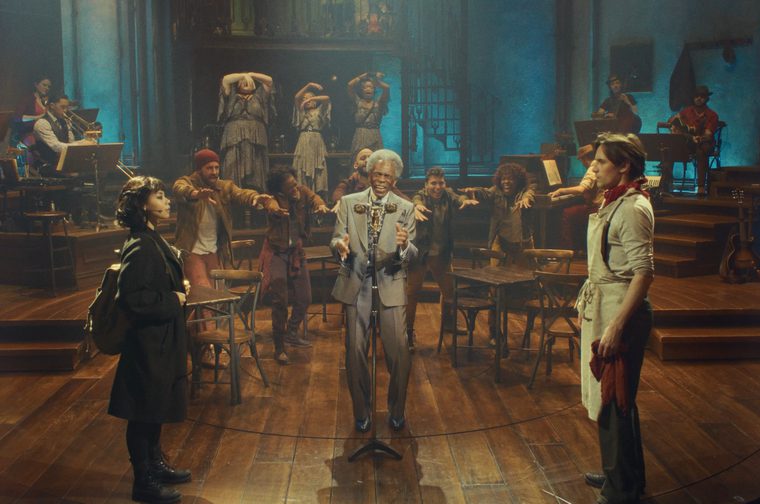 Production photo from the musical Hadestown, showing the full ensemble on a wooden stage set designed like a tavern, with a live band positioned on platforms to either side. In the center, Hermes sings into a vintage microphone wearing a light gray suit, gesturing toward the audience. To his left, Eurydice stands alone in a black coat with a backpack, looking toward him. To his right, Orpheus stands in a cream-colored work shirt and apron with a red cloth in his hand, gazing back at Eurydice across the circle. Behind Hermes, the ensemble of workers reaches out with extended arms, while three women in patterned dresses pose dramatically upstage with their arms raised. The scene is lit in warm amber tones downstage and cool blue light upstage.