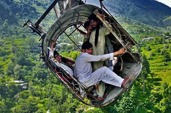 A group of men and boys crowd inside a round metal basket suspended high above a lush green mountain valley, riding an improvised zip-line or cable crossing. One man holds a phone to his ear while others grip the basket's rim; terraced hillsides and a distant village are visible far below.