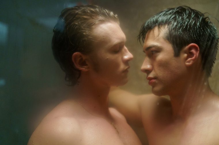 Two young men face each other in close proximity in a steamy, dimly lit shower room. One has light blonde hair and the other has dark hair, both wet. The atmospheric scene is shot with a shallow depth of field, creating a hazy, tense mood.