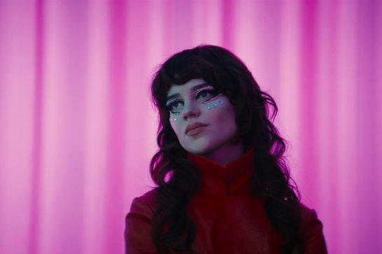 A young white woman with long dark wavy hair and bangs gazes upward with a wistful expression, lit in pink and magenta tones. She wears striking eye makeup with rhinestone streaks below her eyes and small teal teardrop-shaped gems trailing down her cheek like glittering tears. She's dressed in a red high-collared top, set against a backdrop of vertically draped pink curtains.