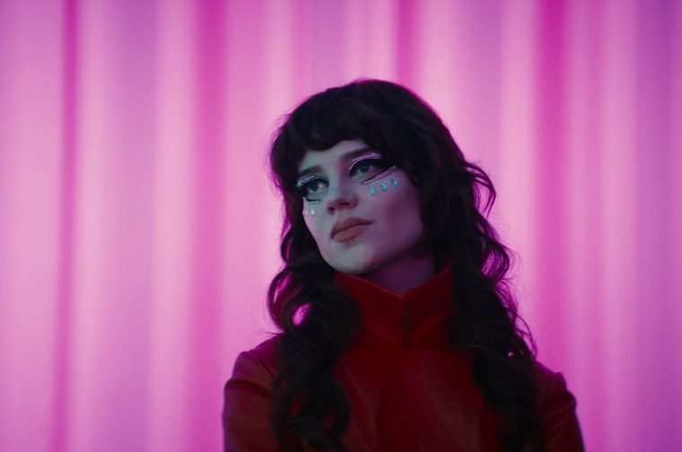 A young white woman with long dark wavy hair and bangs gazes upward with a wistful expression, lit in pink and magenta tones. She wears striking eye makeup with rhinestone streaks below her eyes and small teal teardrop-shaped gems trailing down her cheek like glittering tears. She's dressed in a red high-collared top, set against a backdrop of vertically draped pink curtains.
