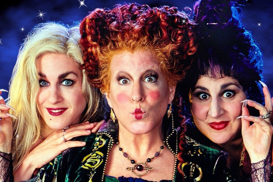 Three witches pose together against a starry night-sky background. The one on the left has long blonde hair and pale makeup; the one in the center has a tall pouf of curly red hair, exaggerated blush, and a pursed-lip expression; the one on the right has dark curly hair, dramatic wide eyes, and long pointed fingernails raised beside her face. All three wear ornate green and purple velvet costumes with gold embroidery and beaded jewelry.
