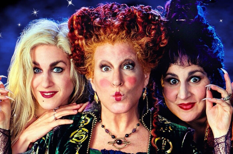 Three witches pose together against a starry night-sky background. The one on the left has long blonde hair and pale makeup; the one in the center has a tall pouf of curly red hair, exaggerated blush, and a pursed-lip expression; the one on the right has dark curly hair, dramatic wide eyes, and long pointed fingernails raised beside her face. All three wear ornate green and purple velvet costumes with gold embroidery and beaded jewelry.