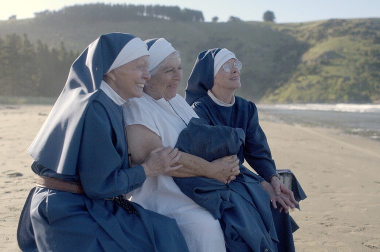 Three elderly nuns in blue and white habits sit together on a beach, smiling and laughing as they look off to the side. Green coastal hills and pine trees rise in the background under a hazy sky.