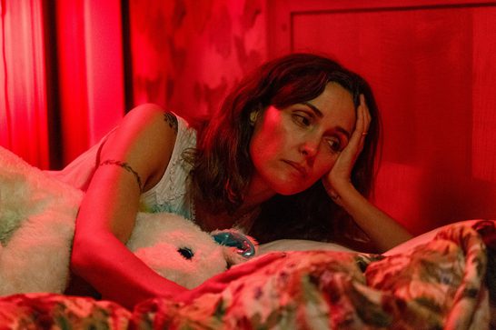 A sombre looking woman in her forties with long brunette hair (Rose Byrne) lies on a bed, her head propped up by her left hand. She is hugging a white stuffed plushie and the image is lit up in stark red.