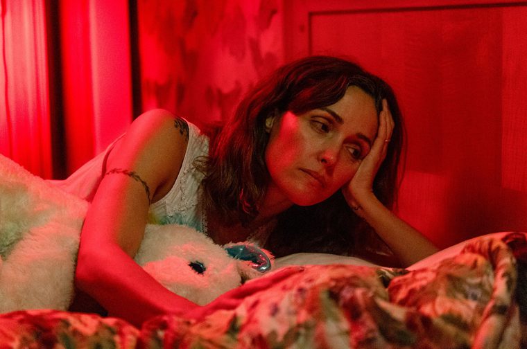 A sombre looking woman in her forties with long brunette hair (Rose Byrne) lies on a bed, her head propped up by her left hand. She is hugging a white stuffed plushie and the image is lit up in stark red.