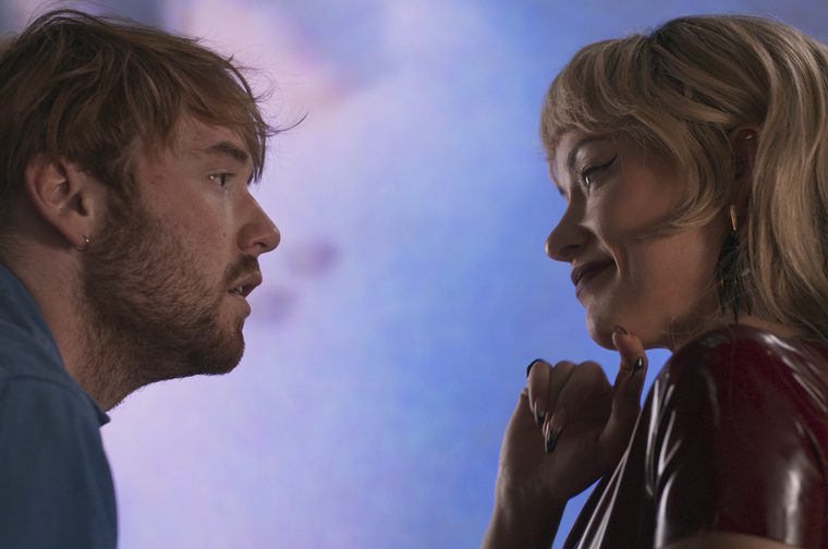 A close-up profile shot of two people facing each other in intimate, dim lighting. On the left, a young-ish man with tousled reddish-brown hair, an earring, and a beard, wearing a blue collared shirt, gazes intently at the woman across from him. On the right, a fortysomething woman with wavy blonde hair, winged eyeliner, dark lipstick, and dark manicured nails wearing a burgundy latex or vinyl garment touches her fingers to her chin as she looks back at him with a slight smile. The background is a soft, blurred gradient of blue and lavender tones, suggesting a nightclub or party atmosphere.