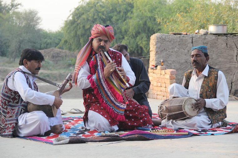 Three South Asian musicians perform outdoors on a colorful patterned rug. The central figure wears a vivid red embroidered vest with tasseled fringe and a red-and-purple striped turban, playing a double-piped wind instrument (likely a satara/algoza). To the left, a man in a white kurta and embroidered vest plays a stringed instrument resting on his lap. To the right, a man in a blue cap and patterned vest plays a dhol-style hand drum. A fourth partially visible person sits behind them. The backdrop shows a mud-brick structure and dense green trees under a hazy sky.