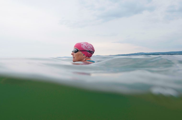 An open water swimming shot taken at the waterline. A female swimmer in her fifties is wearing a pink swim cap and goggles, with just her head visible above the calm, glassy water.