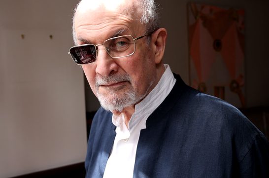A man with white hair and a beard wears glasses with one lens darkened, looking directly at the camera with a serious expression. He's dressed in a white collarless shirt under a navy blue linen jacket, standing indoors with an abstract orange and white artwork visible in the blurred background.