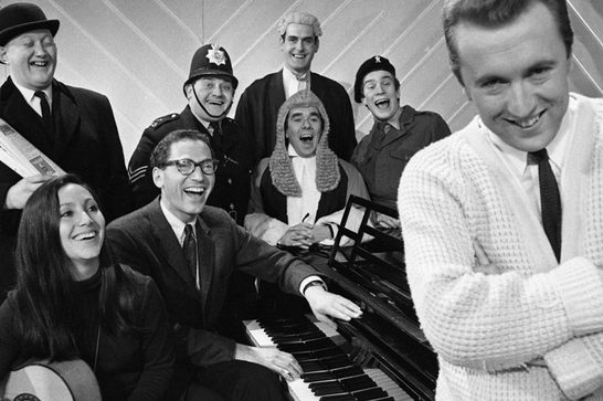 Black-and-white publicity photo of a group of people in comic costumes (bowler hat, policeman, judge, barrister-style wig) gathered around a piano, laughing, with a man in a white cardigan standing in the foreground with arms crossed and smiling at the camera.