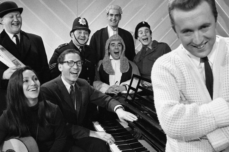 Black-and-white publicity photo of a group of people in comic costumes (bowler hat, policeman, judge, barrister-style wig) gathered around a piano, laughing, with a man in a white cardigan standing in the foreground with arms crossed and smiling at the camera.