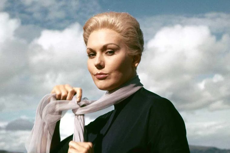 We see a woman (actress Kim Novak) with platinum blonde hair swept up in an elegant updo, wearing a dark top and holding a sheer lavender/grey scarf. She has a cool, composed expression and is photographed against a dramatic cloudy sky, giving the image a moody, cinematic quality.