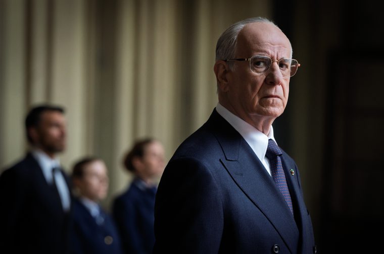The image shows an elderly man in a dark navy double-breasted suit with a patterned tie and gold-framed glasses. He has a stern, serious expression and silver hair. He looks at the viewer and we also see three blurred figures in the background.