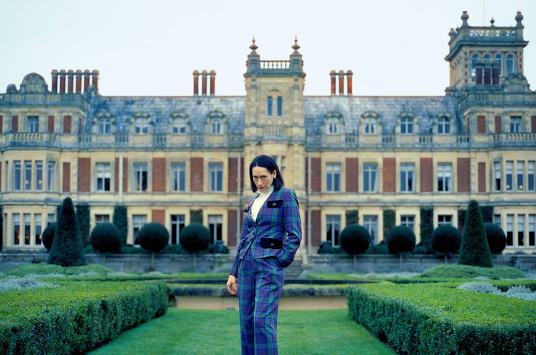A dark-haired woman wearing a blue and green tartan three-piece suit stands alone in the manicured formal gardens of a large Victorian-era country house. She looks downward with a composed expression. Behind her, the stone and red-brick mansion features ornate architectural details, tall chimneys, and rows of large windows. The symmetrical garden is lined with clipped topiary shrubs and hedges. The overcast sky gives the scene a cool, moody atmosphere.