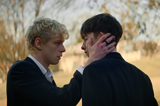 Two young men in dark suits stand close, face to face, in a tree-lined outdoor setting at golden hour; the curly-haired blond man cups the back of the dark-haired man's head with his hand, a small scratch visible on the dark-haired man's cheek, as they hold an intense, emotional gaze.
