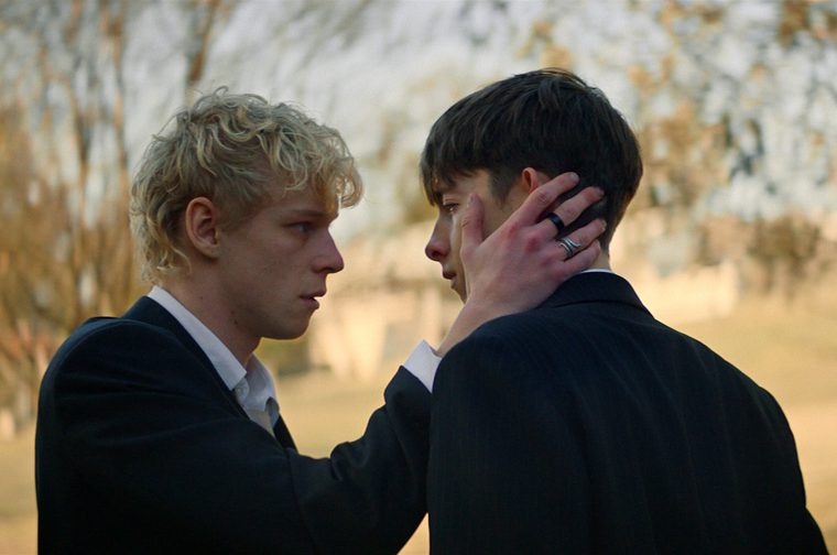 Two young men in dark suits stand close, face to face, in a tree-lined outdoor setting at golden hour; the curly-haired blond man cups the back of the dark-haired man's head with his hand, a small scratch visible on the dark-haired man's cheek, as they hold an intense, emotional gaze.