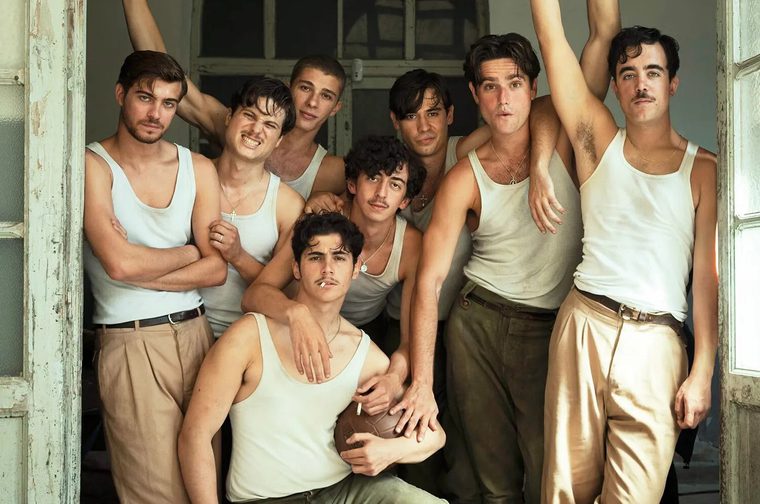 A group portrait of eight young men crowded into a doorway with chipped, pale green paint. They wear white tank tops and loose, high-waisted trousers in khaki and olive, several with leather belts and thin chain necklaces. Their poses are relaxed and playful: one crouches in front holding a vintage leather football with a cigarette in his mouth, one grimaces comically at the camera, and others lean on each other or raise an arm overhead.