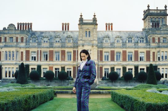 A dark-haired woman wearing a blue and green tartan three-piece suit stands alone in the manicured formal gardens of a large Victorian-era country house. She looks downward with a composed expression. Behind her, the stone and red-brick mansion features ornate architectural details, tall chimneys, and rows of large windows. The symmetrical garden is lined with clipped topiary shrubs and hedges. The overcast sky gives the scene a cool, moody atmosphere.