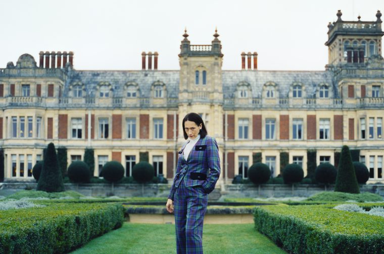 A dark-haired woman wearing a blue and green tartan three-piece suit stands alone in the manicured formal gardens of a large Victorian-era country house. She looks downward with a composed expression. Behind her, the stone and red-brick mansion features ornate architectural details, tall chimneys, and rows of large windows. The symmetrical garden is lined with clipped topiary shrubs and hedges. The overcast sky gives the scene a cool, moody atmosphere.