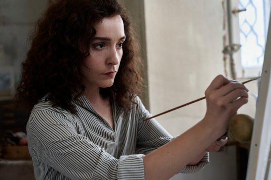 A young woman with curly dark brown hair concentrates intently as she paints on a canvas, holding a thin paintbrush. She wears a striped button-up shirt and is illuminated by natural light from a window in the background. The setting appears to be an artist's studio.