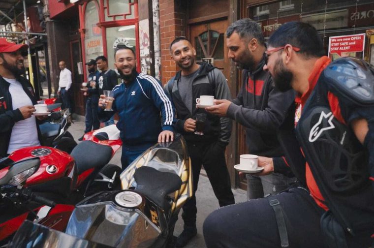 alt text for this image19:14Claude responded: A group of men gathered on a London sA group of South Asian men are gathered on a London street beside parked motorcycles, smiling and chatting while holding cups of tea or coffee. The scene has a casual, social atmosphere, with brick-fronted shops visible in the background.