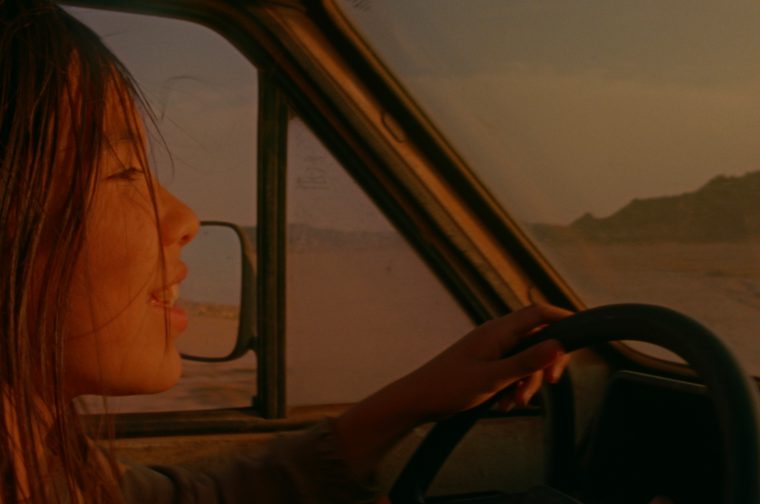 A young girl with windswept hair drives a vehicle at golden hour, her face lit warmly as she looks ahead with mouth open mid-speech or laughter. The side mirror and window frame are visible, with a hazy desert or coastal landscape and distant hills stretching outside.
