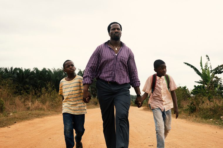A Black man walks hand-in-hand with two young Black boys along a red dirt road in what appears to be a rural African setting. The composition is striking, with the man positioned centrally and slightly elevated in the frame, wearing a purple striped shirt, while the boys flank him on either side - one in an orange striped shirt and the other in a pink shirt with a backpack. The landscape around them features vegetation including banana plants and other greenery, with an overcast sky above. The dirt road stretches ahead of them, creating a sense of journey or purposeful movement forward.
