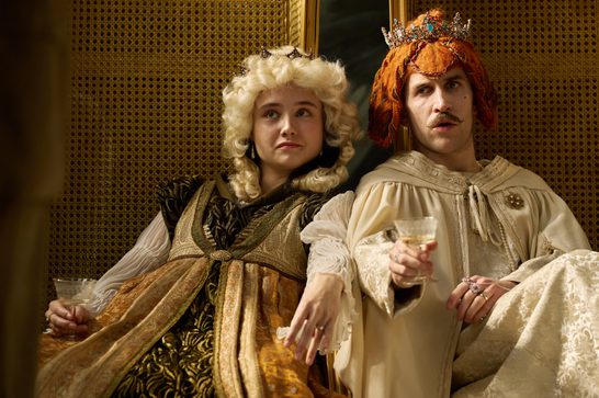 Two performers in elaborate period costume sit together holding wine glasses — one wearing a voluminous blonde baroque wig and ornate gold brocade gown with a small crown, the other in cream robes with a bold auburn wig topped by a jewelled crown and a styled moustache, both gazing off with theatrical expressions against a woven cane backdrop.